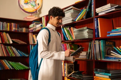 البحث عن معلومات مفيدة وهامة، دمج التقنية والتكنولوجيا مع التعلم، فتى عربي خليجي سعودي يرتدي الثوب التقليدي وحقيبة الظهر يحمل بيده جهازالتابلت، متابعة مقاطع ثقافية وحضور دورات تعليمية الكترونية