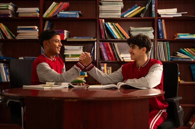 Getting to know each other and forming successful social relationships, education in the Kingdom of Saudi Arabia, completing school assignments, two Arab Gulf Saudi students wearing school uniforms sitting in the school library with shelves full of books behind them shaking hands, enjoying reading school books.