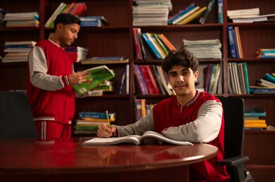 التعليم في المملكة العربية السعودية، تأدية الواجبات الدراسية، طالبان عربيان خليجيان سعوديان يرتديان الزي المدرسي يجلسان في المكتبة المدرسية وخلفهم رفوف مليئة بالكتب، الاستمتاع بقراءة الكتب المدرسية