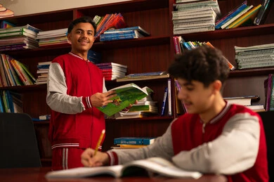 Two Arab Gulf Saudi students are sitting in the school library with shelves full of books behind them, enjoying reading textbooks, doing homework, and education in the Kingdom of Saudi Arabia.