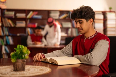 التعلم الذاتي خلال أوقات الفراغ، تصوير مقرب لطالب يقوم بقراءة كتاب، معلم عربي خليجي سعودي يرتدي الثوب التقليدي والشماغ  يقوم بمناقشة الدروس مع تلميذه داخل المكتبة المدرسية، حوارات ونقاشات بين الاستاذة و الطلبة