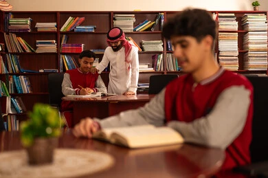 تصوير مقرب لطالب يقوم بقراءة كتاب، قضاء وقت الفراغ بالتعلم والتثقف الذاتي ،  معلم عربي خليجي سعودي يرتدي الثوب التقليدي والشماغ  يقوم بمناقشة الدروس مع تلميذه داخل المكتبة المدرسية، حوارات ونقاشات بين الاستاذة و الطلبة