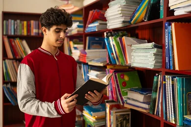 الشغف بالمعرفة والاستطلاع، زيارة مكتبة المدرسة، طالب عربي خليجي سعودي يرتدي الزي المدرسي يقف امام مكتبة خشبية يحمل بيده كتاب، تنمية مهارة القراءة للطلاب، التعليم والدراسة في السعودية ، قضاء وقت الفراغ بالتعلم والتثقف الذاتي