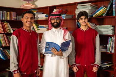 قضاء وقت ممتع ، ممارسة الهوايات المفضلة ، معلم عربي خليجي سعودي يرتدي الثوب الابيض و الشماغ يقف بجوار مكتبة خشبية في المكتبة المدرسية يقف برفقة طلابة يمسك بيدية الكتاب، الاستمتاع بقراءة الكتب المدرسية، التعليم في المملكة العربية السعودية