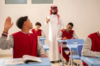 طالب عربي خليجي سعودي يرتدي الزي المدرسي يرفع يديه للإجابة، تعابير وايماءات الحماس لمعرفة الجواب الصحيح، التفاعل والمشاركة داخل الفصل الدراسي، التربية والتعليم بالمناهج المقررة للطلاب في المملكة العربية السعودية