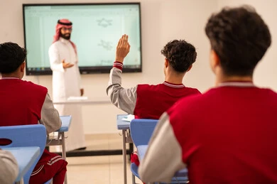 تعابير وايماءات الحماس لمعرفة الجواب الصحيح، طالب عربي خليجي سعودي يرتدي الزي المدرسي يرفع يديه للإجابة، التفاعل والمشاركة داخل الفصل الدراسي، التربية والتعليم بالمناهج المقررة للطلاب في المملكة العربية السعودية