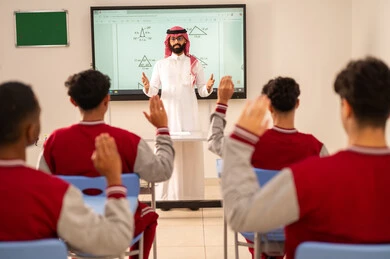 طالب عربي خليجي سعودي يرتدي الزي المدرسي يرفع يديه للإجابة، تعابير وايماءات الحماس لمعرفة الجواب الصحيح، التفاعل والمشاركة داخل الفصل الدراسي، التربية والتعليم بالمناهج المقررة للطلاب في المملكة العربية السعودية