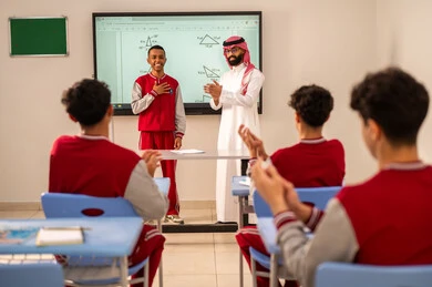 The development of teaching methods in the schools of the Kingdom, honoring a diligent student, hand expressions of excellence and superiority, gestures of joy and happiness, benefiting from advanced technologies in learning and studying, developed curricula and e-learning, Arab Gulf Saudi students sitting in their seats with a Saudi teacher using an electronic board in front of them.