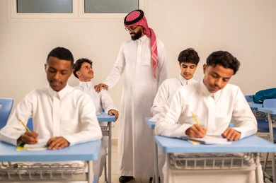معلم عربي خليجي سعودي يرتدي الثوب التقليدي والشماغ يقوم بمناقشة الدروس مع طلابه، حوارات ودية بين الاستاذ والطلاب، المشاركة الفعالة للطلاب مع المدرسين، مجموعة من طلاب عربيين خليجيين سعوديين يرتدون الزي المدرسي يقومون بتأديه الواجبات ، جودة التعليم في السعودية، التربية والتعليم بالمناهج المقررة للتلاميذ