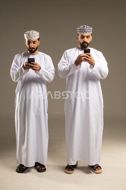 استعمال خدمات الهاتف المحمول, بورتريه لرجلين عربيين خليجيين عمانيين يرتديان الدشداشة و الكمة يستخدمان الموبايل, الإستمتاع مع الأصدقاء, ايماءات الاندماج و التركيز, خلفية رمادية