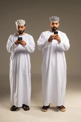 Using mobile phone services, portrait of two Omani Gulf Arab men wearing dishdashas and kumas using mobile phones, enjoying themselves with friends, gestures of engagement and focus, gray background