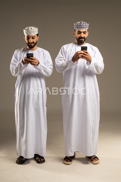 استعمال تطبيقات الهاتف المحمول, بورتريه لرجلين عربيين خليجيين عمانيين يرتديان الدشداشة و الكمة يستخدمان الموبايل, الإستمتاع مع الأصدقاء, ايماءات السعادة و السرور, خلفية رمادية
