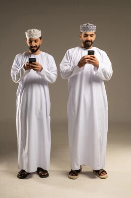 استعمال تطبيقات الهاتف المحمول, بورتريه لرجلين عربيين خليجيين عمانيين يرتديان الدشداشة و الكمة يستخدمان الموبايل, الإستمتاع مع الأصدقاء, ايماءات السعادة و السرور, خلفية رمادية