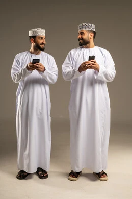 إرسال رسالة نصية عبر الجوال, بورتريه لرجلين عربيين خليجيين عمانيين يرتديان الدشداشة و الكمة يستخدمان الموبايل, الإستمتاع مع الأصدقاء, ايماءات السعادة و السرور, خلفية رمادية