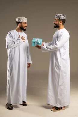 مفهوم الشكر و الامتنان، تقديم الهدايا بالاعياد و المناسبات الخاصة،  بورتريه لرجلين عربيين خليجيين عمانيين يرتديان الدشداشة و الكمة يهدي أحدهما الاخر هدية, خلفية رمادية
