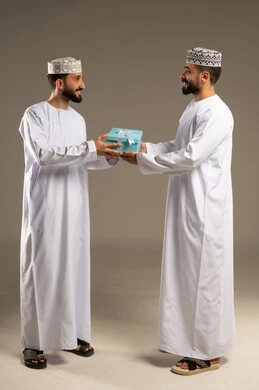 التعبير عن الحب و التقدير, تقديم الهدايا بالاعياد و المناسبات الخاصة، بورتريه لرجلين عربيين خليجيين عمانيين يرتديان الدشداشة و الكمة يهدي أحدهما الاخر هدية, خلفية رمادية