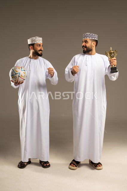 مشاركة للحظة الفوز, مشاهدة و متابعة مباراة كرة القدم، بورتريه لرجلين عربيين خليجيين عمانيين يرتديان الدشداشة و الكمة و يمسك أحدهما بالكرة و الاخر يحمل كأس ذهبي و ينظران لبعضهما, خلفية رمادية