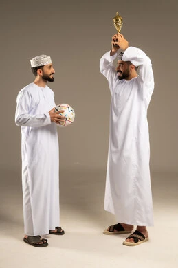 أجواء الاحتفال و السعادة, مشاهدة مباراة كرة القدم، بورتريه لرجلين عربيين خليجيين عمانيين يرتديان الدشداشة و الكمة و يمسك أحدهما بالكرة و الاخر يحمل كأس ذهبي و يرفعه عاليا, خلفية رمادية