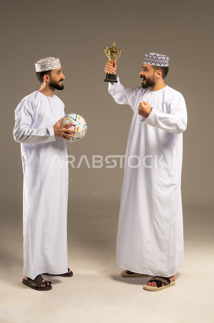 تعبير يدل على الوصول إلى الهدف, مشاهدة و متابعة مباراة كرة القدم، بورتريه لرجلين عربيين خليجيين عمانيين يرتديان الدشداشة و الكمة و يمسك أحدهما بالكرة و الاخر يحمل كأس ذهبي, خلفية رمادية