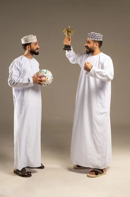 An expression signifying the achievement of a goal; watching and following a football match; a portrait of two Omani Gulf Arab men wearing dishdashas and kumas, one holding a ball and the other a golden cup; gray background.