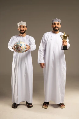 Following a major sporting event, watching a football match, a portrait of two Omani Gulf Arab men wearing dishdashas and kumas, one holding a ball and the other a golden cup, gray background.