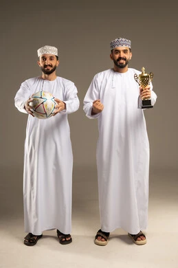 روح الفوز و التشجيع الجماعي, مشاهدة و متابعة مباراة كرة القدم، بورتريه لرجلين عربيين خليجيين عمانيين يرتديان الدشداشة و الكمة و يمسك أحدهما بالكرة و الاخر يحمل كأس ذهبي, خلفية رمادية