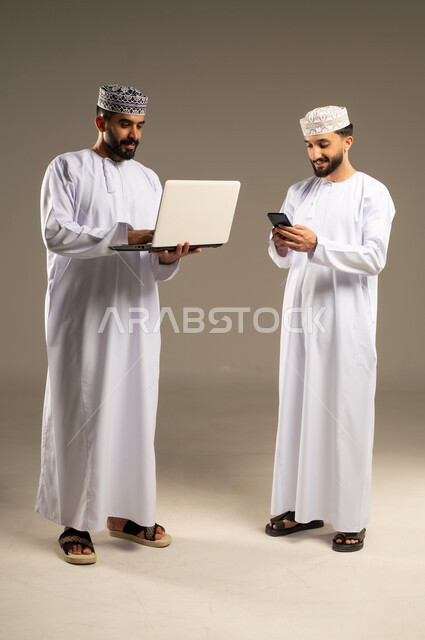 Utilizing modern technology, managing daily work tasks, a portrait of two Omani Gulf Arab men wearing dishdashas and kumas using a laptop and mobile phone, gray background