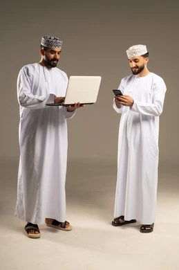 Utilizing modern technology, managing daily work tasks, a portrait of two Omani Gulf Arab men wearing dishdashas and kumas using a laptop and mobile phone, gray background