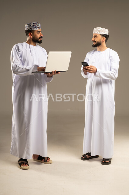 ادارة مهام العمل اليومية, مفهوم التطور و التكنولوجيا، بورتريه لرجلين عربيين خليجيين عمانيين يرتديان الدشداشة و الكمة يستخدمان اللابتوب و الجوال, خلفية رمادية