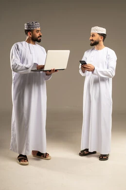 ادارة مهام العمل اليومية, مفهوم التطور و التكنولوجيا، بورتريه لرجلين عربيين خليجيين عمانيين يرتديان الدشداشة و الكمة يستخدمان اللابتوب و الجوال, خلفية رمادية