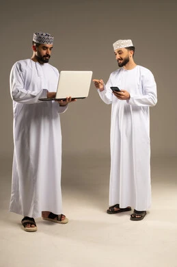 The development of modern technology and techniques, pointing with the index finger at a laptop, a portrait of two Omani Gulf Arab men wearing dishdashas and kumas using a laptop and mobile phone, gray background