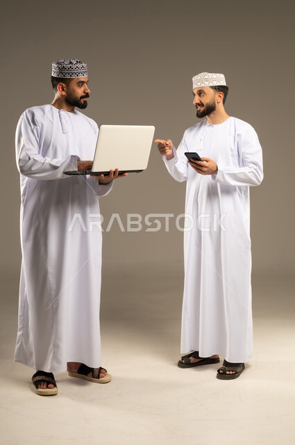 استخدام التكنولوجيا في العمل أونلاين، الاشارة باصبع السبابة الى الحاسوب المحمول, بورتريه لرجلين عربيين خليجيين عمانيين يرتديان الدشداشة و الكمة يستخدمان اللابتوب و الجوال, خلفية رمادية