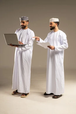 الاشارة باصبع السبابة الى الحاسوب المحمول, بورتريه لرجلين عربيين خليجيين عمانيين يرتديان الدشداشة و الكمة يستخدمان اللابتوب و الجوال, خلفية رمادية