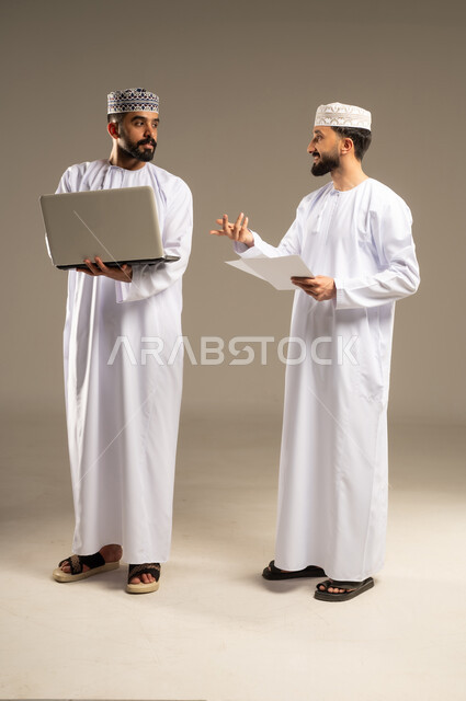 The role of modern technology in the information search process; a portrait of two Omani Gulf Arab men wearing dishdashas and kumas, one using a laptop and the other holding a white sheet of paper and inquiring about something, gray background.