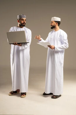 The role of modern technology in the information search process; a portrait of two Omani Gulf Arab men wearing dishdashas and kumas, one using a laptop and the other holding a white sheet of paper and inquiring about something, gray background.