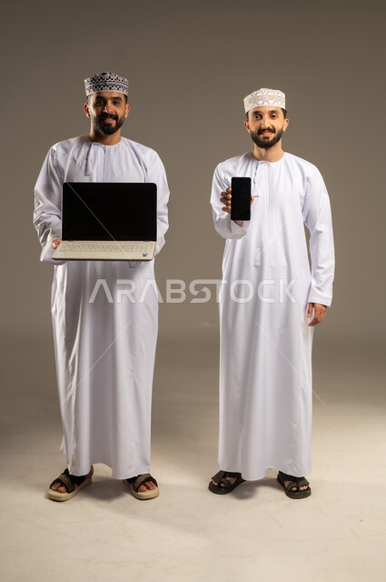Collaboration between friends to accomplish tasks, displaying a blank black screen, portrait of two smiling Omani Gulf Arab men wearing dishdashas and kumas, using a laptop and mobile phone, gray background