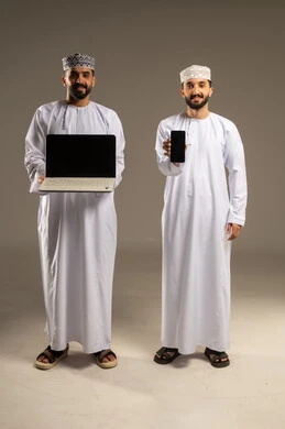 التعاون بين الأصدقاء لإنجاز المهام، عرض شاشة سوداء فارغة,  بورتريه لرجلين عربيين خليجيين عمانيين يرتديان الدشداشة و الكمة مبتسمان يستخدمان اللابتوب و الجوال, خلفية رمادية