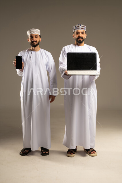 Displayed on a blank black screen via phone and laptop, a portrait of two smiling Omani Gulf Arab men wearing dishdashas and kumas, using a laptop and mobile phone, against a gray background.