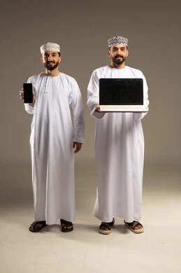 عرض شاشة سوداء فارغة عبر الهاتف و الحاسوب المحمول، بورتريه لرجلين عربيين خليجيين عمانيين يرتديان الدشداشة و الكمة مبتسمان يستخدمان اللابتوب و الجوال, خلفية رمادية