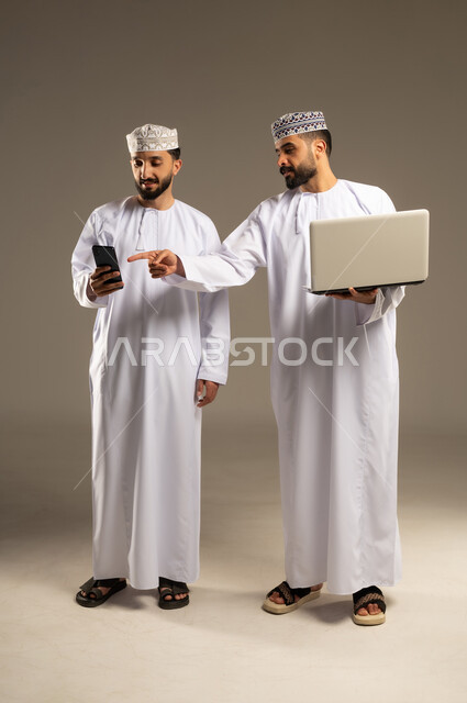 مفهوم التطور و التكنولوجية، العمل عن بعد، بورتريه لرجلين عربيين خليجيين عمانيين يرتديان الدشداشة و الكمة يستخدم أحدهما اللابتوب و يشير باصبع السبابة الى الهاتف, خلفية رمادية