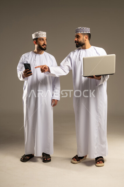 Remote work, the concept of development and technology, a portrait of two Omani Gulf Arab men wearing dishdashas and kumas, one of whom is using a laptop and pointing his index finger at a phone, gray background