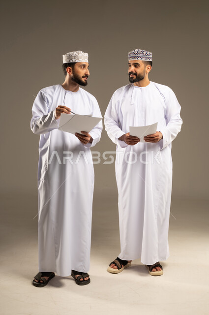 تقسيم و توزيع المهام، الوحده و العمل الجماعي، بورتريه لرجلين عربيين خليجيين عمانيين يرتديان الدشداشة و الكمة يحملان اوراق بيضاء, خلفية رمادية