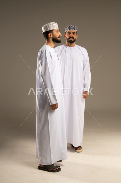 العناية والإهتمام بالمظهر, منظر جانبي لرجل عربي خليجي عماني يرتدي الدشداشة و الكمة بإيماءات وجه و يدين مختلفة يقوم بجلسة تصوير، خلفية لشاب ينظر الى الكاميرا بسرور, تعابير الثقة بالنفس