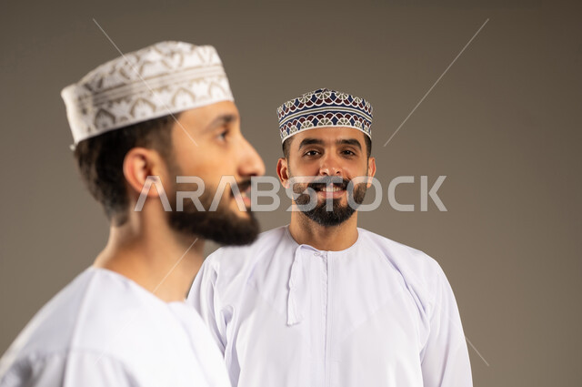 تعبير يدل على البشاشة و السعادة, منظر جانبي لرجل عربي خليجي عماني يرتدي الدشداشة و الكمة بإيماءات وجه و يدين مختلفة يقوم بجلسة تصوير، خلفية لشاب ينظر الى الكاميرا بسرور, تعابير الثقة بالنفس