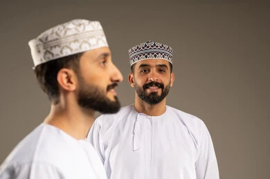 تعبير يدل على البشاشة و السعادة, منظر جانبي لرجل عربي خليجي عماني يرتدي الدشداشة و الكمة بإيماءات وجه و يدين مختلفة يقوم بجلسة تصوير، خلفية لشاب ينظر الى الكاميرا بسرور, تعابير الثقة بالنفس