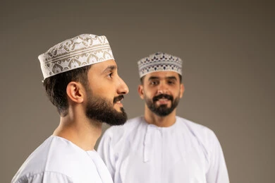 منظر جانبي لرجل عربي خليجي عماني يرتدي الدشداشة و الكمة بإيماءات وجه و يدين مختلفة يقوم بجلسة تصوير، خلفية غير واضحة لشاب ينظر الى الكاميرا بسعادة و سرور, تعابير الثقة بالنفس