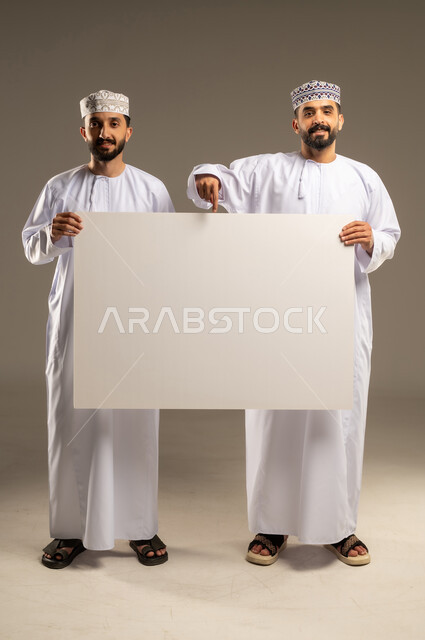 Ensuring clear understanding using the whiteboard, pointing with the index finger to a blank whiteboard, a portrait of two Omani Gulf Arab men wearing dishdashas and kumas, looking happily at the camera, gray background