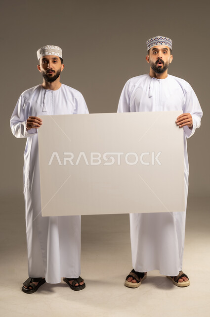 التفاجؤ من شيء ما، استعمال السبورة لتوضيح الاجراءات المتبعة, بورتريه لرجلين عربيين خليجيين عمانيين يرتديان الدشداشة و الكمة و ينظران الى الكاميرا بدهشة, خلفية رمادية