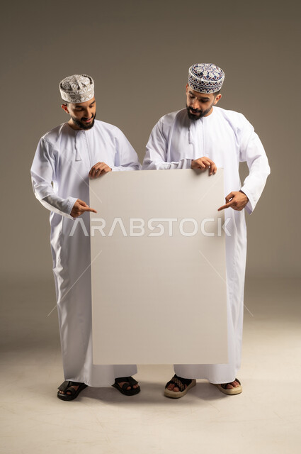 الاشاره بالسبابه الى لوحه بيضاء فارغه, توضيح الاجراءات المتبعة, بورتريه لرجلين عربيين خليجيين عمانيين يرتديان الدشداشة و الكمة و يحملان السبورة, خلفية رمادية