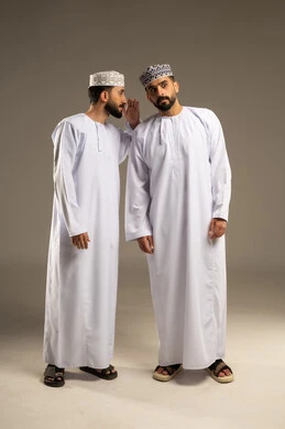 Speaking in a low voice, keeping and preserving secrets, a portrait of two Arab Gulf Omani men wearing the dishdasha and kumma, with one whispering in his friend's ear, gray background.